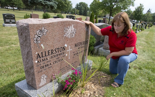 Marg Medeiros kneels next to the gravestone of her mother, Alma Allerston, in St. Peter's Cemetery in London, Ont. on Friday August 14, 2015. Medeiros captured video footage of a woman stealing flowers and mementos from the gravesite on Wednesday after attaching a camera to a nearby tree in hopes of identifying the thief.  Flowers, mementos and hand-made tributes to the woman who had eight children, thirteen grandchildren and more than twenty great grandchildren, have been going missing on a regular basis since Allerston was laid to rest in April.  Craig Glover/The London Free Press/Postmedia Network