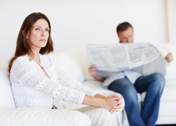 Upset middle aged woman on sofa with man reading newspaper in background